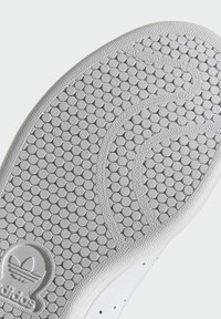 Valge spordijalatsite tallal on ringikujuline tekstuur, millel on sile äär ja logo. Disain rõhutab haarduvust ja stabiilsust.