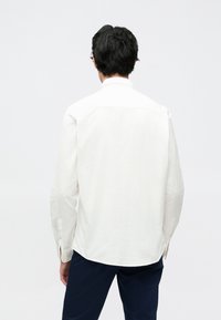 Bela majica z dolgimi rokavi iz bombaža. Ima klasičen ovratnik, zapete manšete in ukrivljen spodnji rob. Preprosta, gladka tekstura, brez vzorcev.