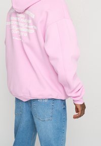 Haut à capuche rose avec du texte imprimé blanc dans le dos, texture douce et coupe décontractée. Assorti à un jean en denim bleu clair avec des détails de logo.