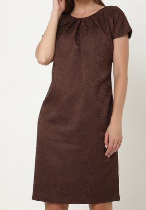 Kobieta w ciemnobrązowej sukience do kolan z krótkim rękawem i subtelną fakturą paisley, pozująca z jedną ręką przy twarzy, a drugą wzdłuż ciała.