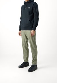 Felpa nera con cappuccio e tasca frontale, abbinata a pantaloni leggeri di colore verde chiaro con texture e sneakers nere, creando un outfit casual.