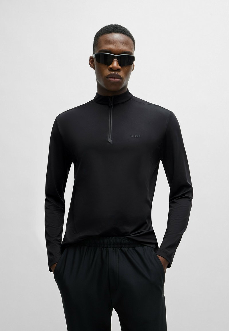 Top negro de manga larga con cremallera, hecho de una tela suave, que presenta un cuello alto y un discreto detalle de logo. Combinado con pantalones negros de cintura elástica.