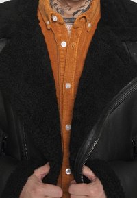 Mani che stringono i risvolti di un giubbotto in pelle shearling nera indossato sopra una camicia a bottoni in corda arancione, con un tatuaggio visibile sul collo.