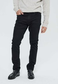 Homme portant un pantalon noir slim fit, des mocassins en cuir noirs et un pull en tricot gris clair avec une main dans la poche, se tenant contre un fond blanc.