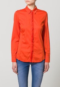 Camicia rossa con bottoni, dotata di una patta anteriore testurizzata e maniche lunghe, realizzata in un tessuto leggero. Abbinata a jeans blu.