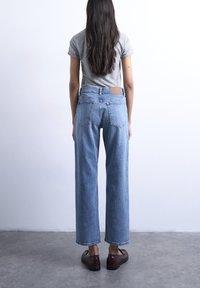 Άτομο με μακριά σκούρα μαλλιά που φοράει γκρι μπλουζάκι, ψηλόμεσο μπλε τζιν και καφέ παπούτσια, στέκεται σε τσιμεντένιο δάπεδο μπροστά σε λευκό τοίχο.