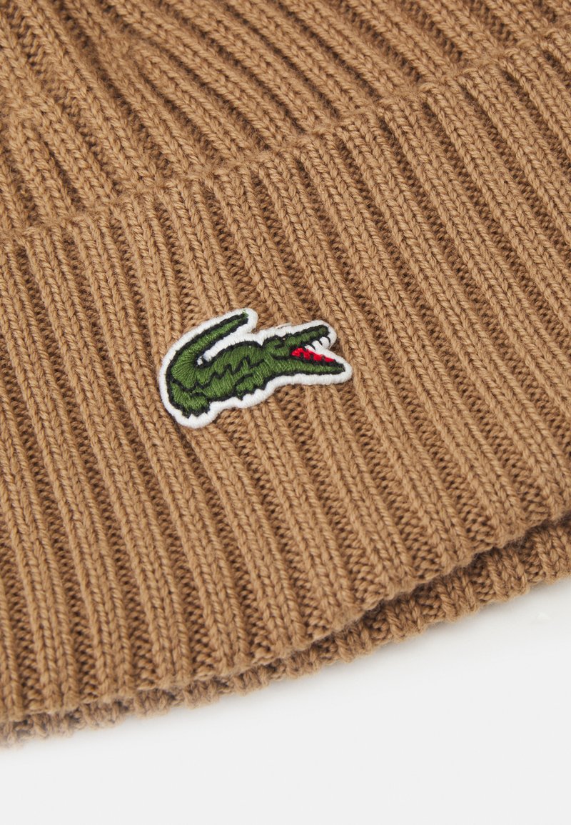 Un bonnet en tricot côtelé marron avec un logo de crocodile brodé en vert et des accents rouges. La texture est douce et extensible.