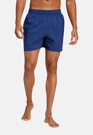 Homme portant un short de bain bleu, debout pieds nus sur fond blanc, montrant le devant et les jambes avec les mains détendues le long du corps.