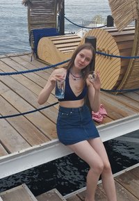 Ein Model in einem marineblauen Bikini und einem Jeansrock hält ein klares Glas mit blauer Flüssigkeit und einem kleinen runden Objekt und sitzt auf einem Holzsteg.