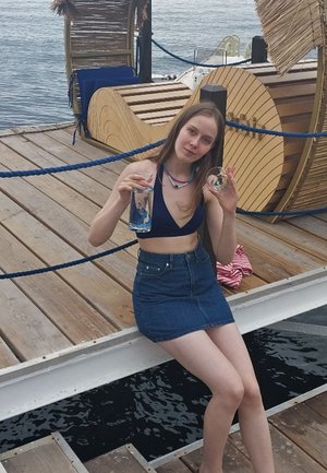 Model in a navy bikini and denim skirt holds a clear glass with blue liquid and a small circular object, seated on a wooden dock.