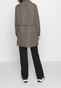 Kaki trenchcoat med en något oversized passform, som har hög krage och knäppte manschetter, tillsammans med svarta byxor och sportskor.