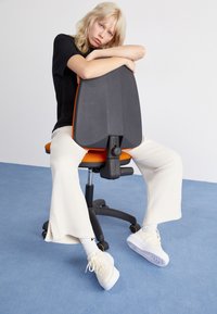 Černá ergonomická kancelářská židle s oranžovými akcenty, elegantní sedadlo a texturované opěradlo. Model má na sobě krémové kalhoty a bílé tenisky.