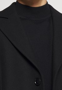 Personne portant un manteau noir boutonné par-dessus un pull à col haut côtelé noir, mettant en valeur la texture du tissu et les détails du revers du manteau.