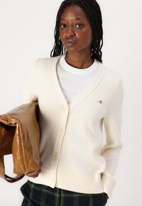 Cardigan color crema con scollo a V, chiusura con bottoni e orlo a coste. Presenta un piccolo logo ricamato. Abbinato a una camicia bianca e pantaloni a quadri.