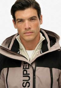 Voorkant van een hooded jack met een zwart en lichtroze kleurenschema, met een opvallend "SUPER" logo, ritsluiting en een gestructureerde kraag.