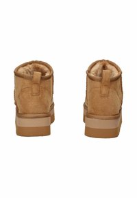 Un paio di stivali alla caviglia in suede marrone chiaro con rivestimento in pelliccia morbida, visibili da dietro, dotati di linguette per indossarli e suole in gomma spessa.