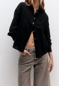 Chemise en lin noire à boutons, avec une coupe décontractée et des manches larges, associée à un jean taille haute gris clair. Tissu texturé visible.