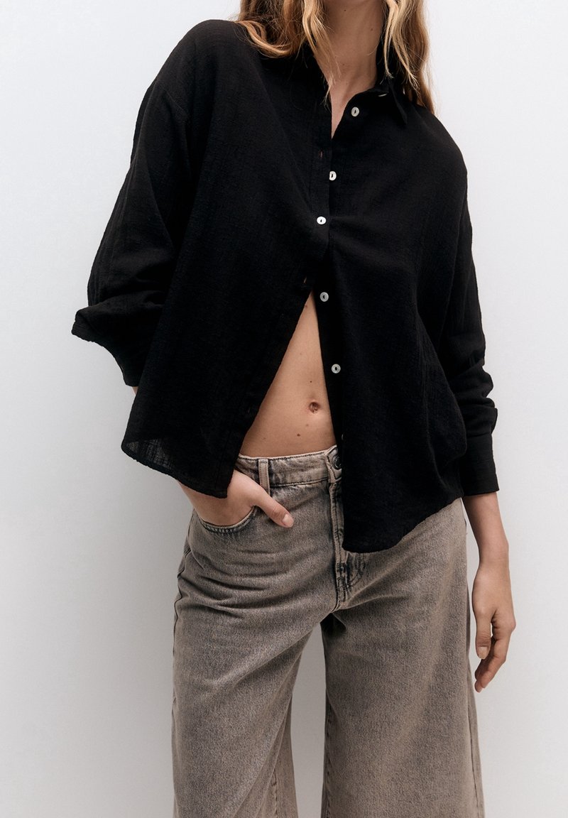 Chemise en lin noire à boutons, avec une coupe décontractée et des manches larges, associée à un jean taille haute gris clair. Tissu texturé visible.