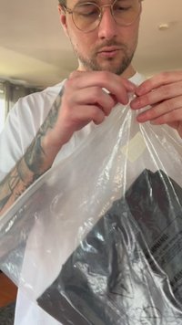 Prozoren plastični paket se odpira, razkrivajoč črno izdelavo z opozorilnimi oznakami. Osredotočite se na teksturo in prosojnost embalaže.