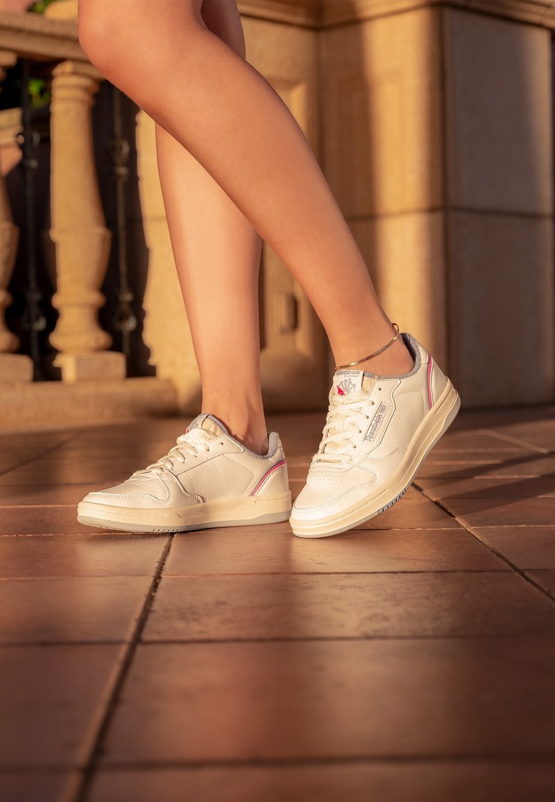 Białe sneakersy z teksturowanej skóry, okrągłym czubkiem, sznurowanym zapięciem i subtelnym różowym akcentem. Lekka podeszwa, płaski profil i minimalne oznakowanie.