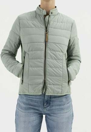 Person trägt eine hellgrüne gesteppte Jacke mit Frontreißverschluss und Reißverschlusstaschen an den Seiten, kombiniert mit blauen Jeans, Hände in den Taschen.