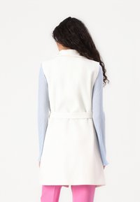 Chaleco blanco sin mangas con un cinturón, con una textura suave y un cuello afilado. Combinado con una camiseta de manga larga azul con relieve y pantalones rosas.