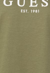 Camiseta verde oliva con texto impreso en blanco que dice "GUESS" y "EST. 1981", confeccionada en un material de algodón suave y ligero.