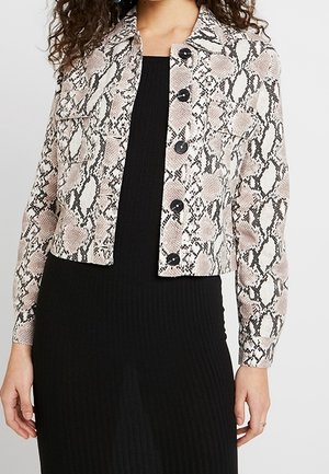 Žena oblečená v krátké bundě se vzorem hadí kůže a knoflíky přes dlouhé černé žebrované šaty, stojící s rukama uvolněně podél těla.