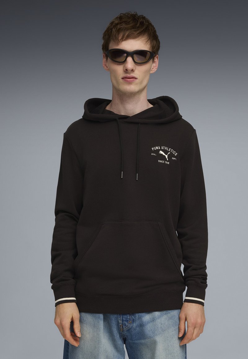 Joven con sudadera negra de Puma Athletics, gafas de sol y vaqueros azul claro, posando frente a un fondo degradado gris.