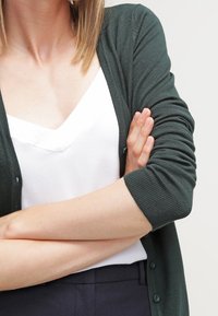 Mörkgrön kardigan med långa ärmar, ribbad struktur och knäppning, lagd över en vit linne, vilket visar en avslappnad stil.