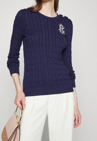 Marinblå kabelstickad tröja med ribbade manschetter och nederkant, utrustad med dekorativa vita knappar på axeln samt en vit logotyp på bröstet.