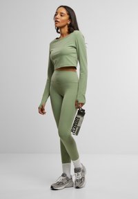 Olive green crop top with long sleeves and high-waisted leggings. Model holds a clear water bottle. Sneakers with grey accents and white socks.