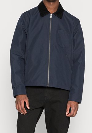 Homme portant une veste zippée bleu marine avec col noir et poche poitrine, associée à un pantalon noir, debout devant un fond clair.