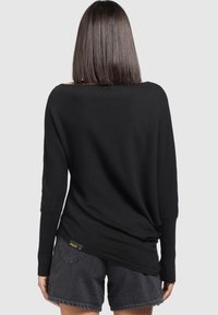 Pull noir surdimensionné avec manches longues et poignets côtelés ; texture douce et coupe décontractée ; étiquette visible en bas à gauche.