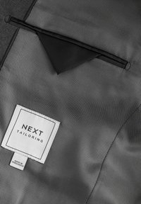 Szary materiał o fakturze zawiera czarną kieszeń oraz białą metkę z napisem "NEXT TAILORING", z mniejszą etykietą informującą "Wyprodukowano w Bangladeszu".