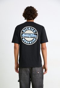 T-shirt en coton noir avec un graphique circulaire bleu et crème au dos, comportant le texte "DICKIES WORKWEAR" et une illustration de camion vintage.
