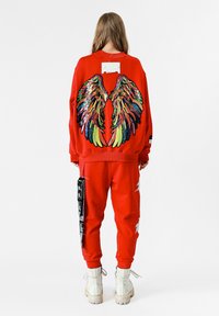 Sudadera roja con diseño de alas de lentejuelas multicolores en la parte posterior, combinada con pantalones rojos a juego que cuentan con una etiqueta gráfica lateral y botas blancas.