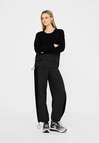 Zwarte trui met lange mouwen en een aansluitend ontwerp, gecombineerd met zwarte wijdvallende broek met zijritsen. Model draagt meerkleurige sportschoenen.