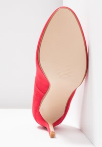 Czerwony but na wysokim obcasie wykonany z miękkiego materiału, z ostro zakończonym noskiem i smukłym obcasem typu stiletto. Podeszwa jest gładka i jasna.