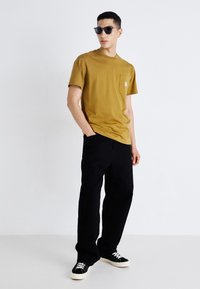 T-shirt giallo senape a maniche corte con taschino sul petto, abbinata a pantaloni neri e sneakers nere con lacci bianchi. Modello che indossa occhiali da sole.