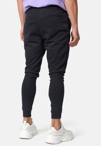 Hombre vistiendo pantalones jogger negros y zapatillas blancas gruesas, de pie con las piernas ligeramente separadas, vista desde atrás sobre fondo blanco.