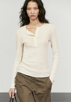 Donna con i capelli lunghi e scuri indossa una maglia henley color crema a maniche lunghe e pantaloni marroni, in piedi con una mano in tasca e l'altra che tiene una borsa.