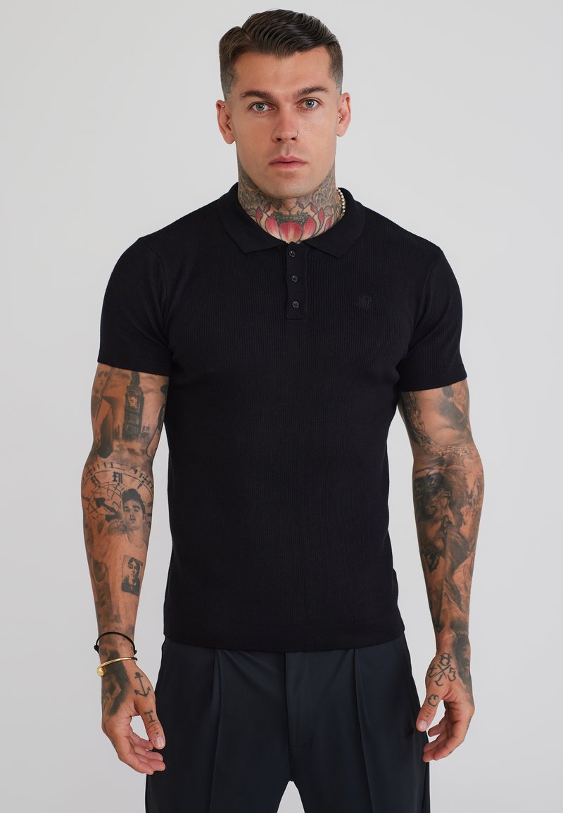 Man with tattoos on neck and arms wearing a fitted black short-sleeve polo shirt and dark pants against a plain background.