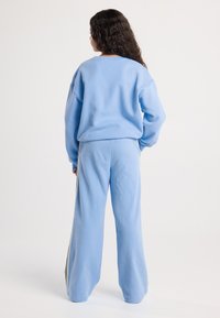 Nainen, jolla on pitkät kiharat hiukset, seisoo selin, päällään löysä vaaleansininen collegepaita ja siihen sopivat leveälahkeiset housut, joissa on sivustripit.