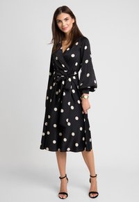 Czarna sukienka o kroju kopertowym w beżowe kropki, z długimi, balonowymi rękawami, paskiem w talii oraz spódnicą do kolan. Gładki materiał o delikatnym połysku.