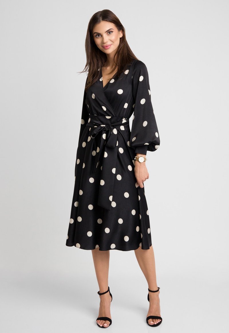 Czarna sukienka o kroju kopertowym w beżowe kropki, z długimi, balonowymi rękawami, paskiem w talii oraz spódnicą do kolan. Gładki materiał o delikatnym połysku.