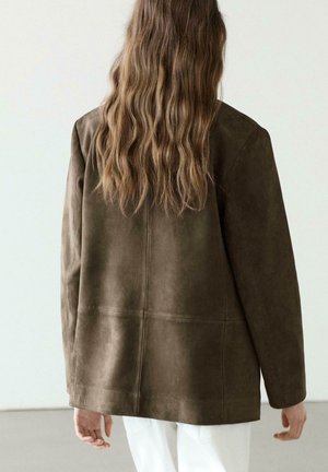 Person mit langen, welligen Haaren, die eine übergroße dunkelbraune Wildlederjacke und weiße Hose trägt, vor einem schlichten hellen Hintergrund stehend.