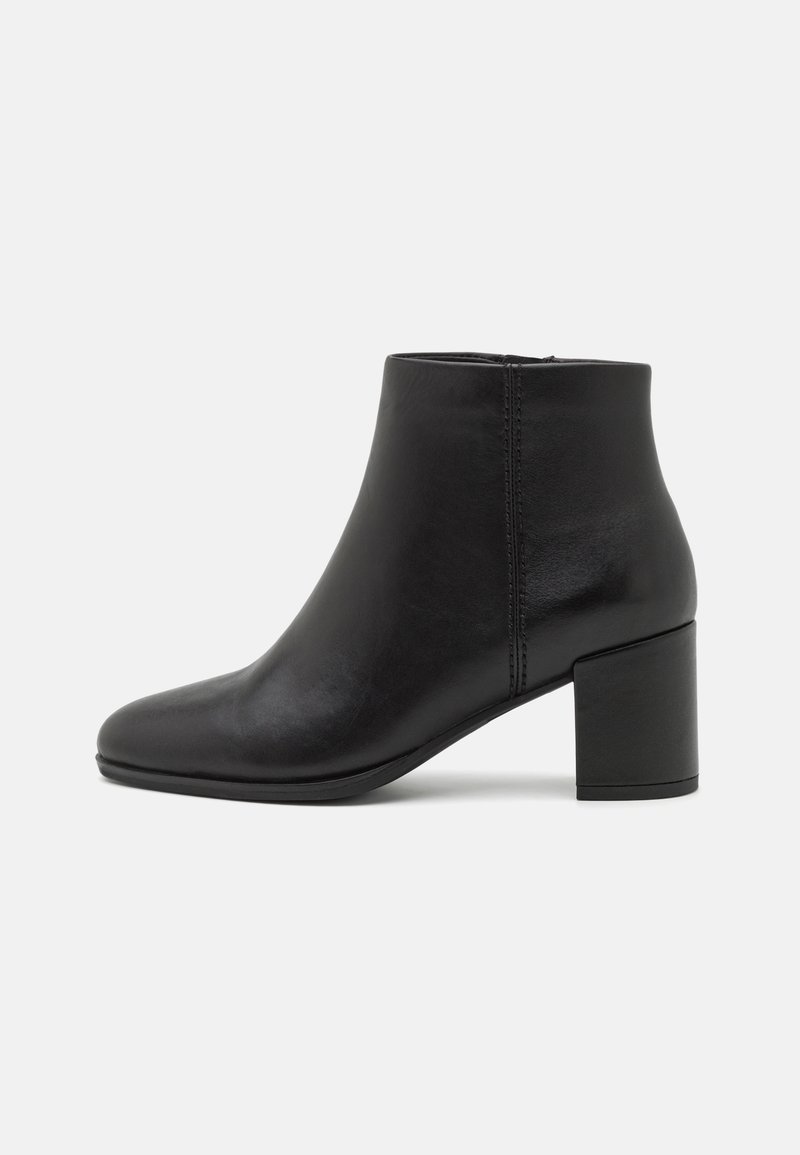 Bottine en cuir noir avec une fermeture éclair latérale et un talon bloc épais, sur un fond gris clair uni.
