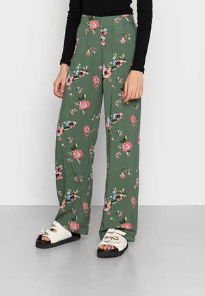 Calças largas florais em tecido verde com flores rosas e azuis. Apresentam um design de cintura alta e um ajuste confortável. Combinadas com sandálias brancas.