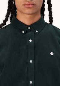 Cămașă butonată din corduroy verde închis, cu guler clasic, butoane albe și un mic logo alb pe partea stângă a pieptului.